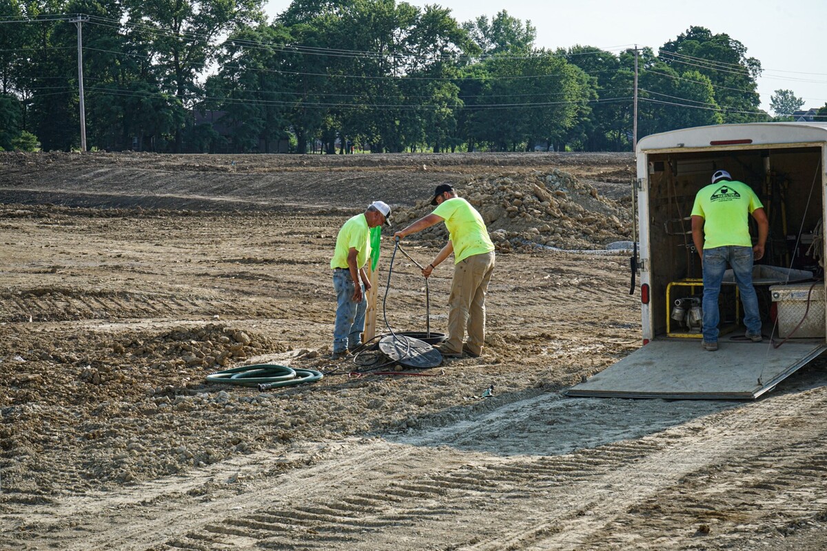 two worker stand over a hole next to them a third worker is retrieving something from a trailer