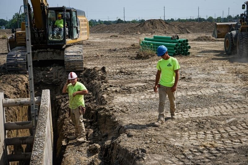 a worker stand in a hole as another watches